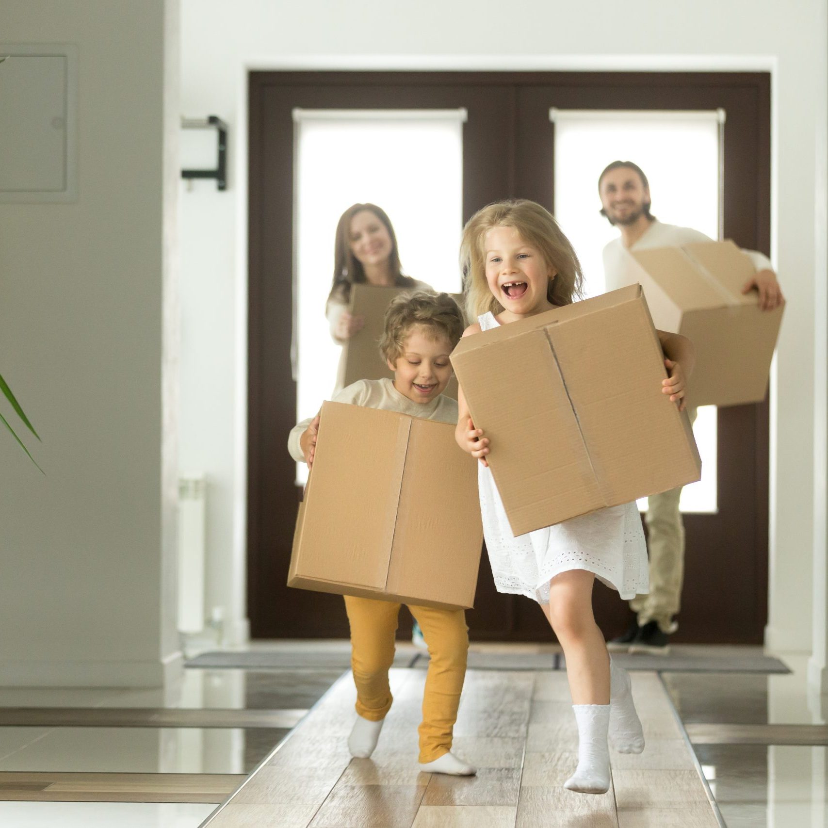 Happy family with kids bought new home, excited children funny girl and boy holding boxes running into big modern house, helping parents with belongings, moving day concept, mortgage and relocation